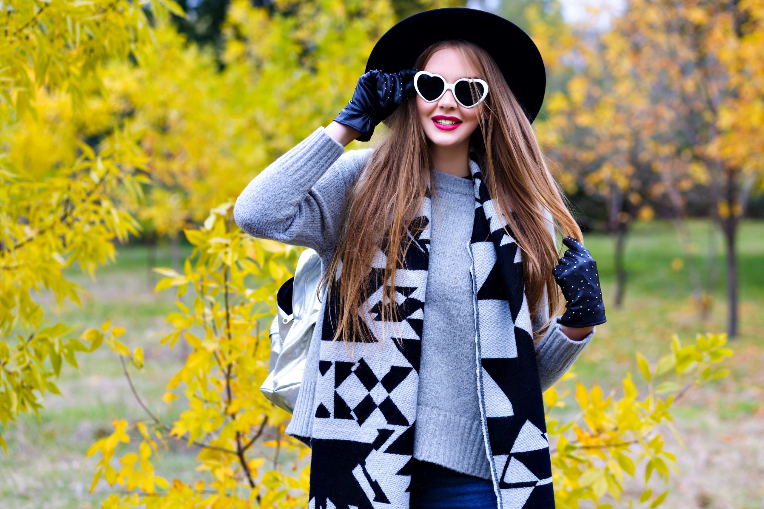 Glad woman wears elegant coat and black hat posing in sunglasses in sunny autumn day. Outdoor portrait of enthusiastic female model in trendy gray sweater walking in park with yellow trees.